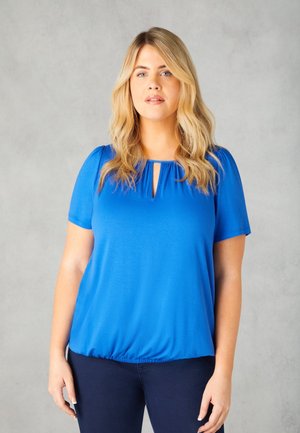 Top blu corto a maniche corte con scollo rotondo e dettaglio a OCCHIO DI BUE. Tessuto morbido ed elasticizzato con orlo arricciato, indossato con pantaloni blu scuro.