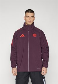 Giacca a vento bordeaux con strisce rosse sulle spalle, chiusura con zip e logo sul petto. Realizzata in materiale leggero e resistente all'acqua.