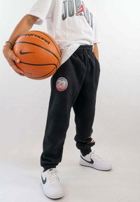 Zwarte joggingbroek met een circulair '23' logo, gecombineerd met een wit t-shirt met de tekst 'JORDAN', en witte Nike sneakers. De persoon houdt een basketbal vast.