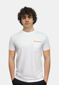 T-shirt bianco a maniche corte con accenti arancioni sulle maniche e nella tasca. Presenta una tasca bianca a contrasto e dettagli nel colletto di colore bianco.