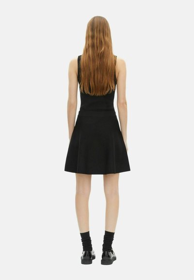 Femme aux longs cheveux bruns portant une robe noire sans manches, des chaussettes noires et des chaussures noires brillantes, se tenant de dos sur un fond blanc.
