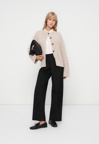Knit beige cardigan with a textured pattern, round button closure, white shirt, black wide-leg trousers, and black slip-on shoes.