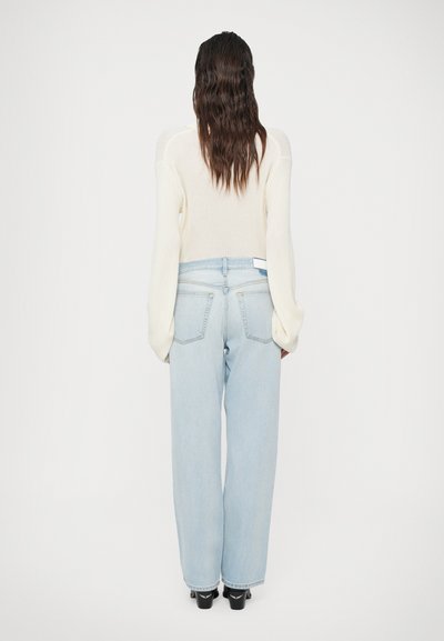 Maglione a coste color crema con maniche lunghe abbinato a jeans a vita alta larghi di colore azzurro chiaro e scarpe nere, visto da dietro.