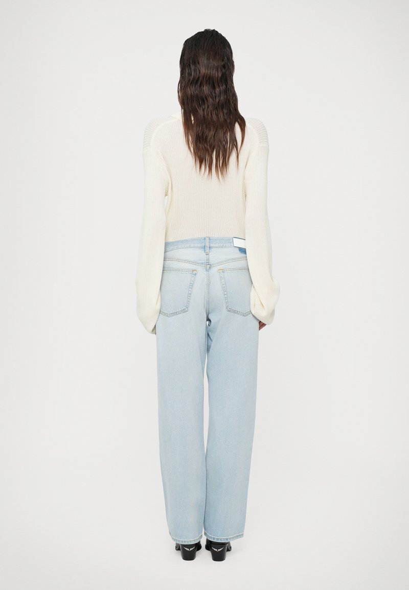 Un pull côtelé de couleur crème à manches longues associé à un jean large taille haute bleu clair et des chaussures noires, vu de dos.