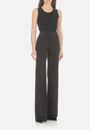 Frau trägt ein schwarzes ärmelloses Oberteil und schwarze, hoch taillierte, weite Hose mit spitzen schwarzen Schuhen, steht vor einem einfarbigen Hintergrund.