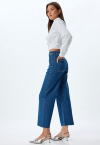 Jeans de mezclilla azul de pierna ancha con cintura alta y dobladillo sin rematar, combinados con una camiseta corta blanca a rayas de manga larga. Sandalias con tacón plateadas.