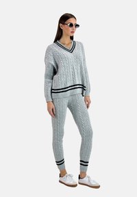 Grå kabelstickad tröja med V-ringning och svart rand i nederkant, i kombination med matchande kabelstickade byxor och vita sneakers. Tillbehören inkluderar runda solglasögon.