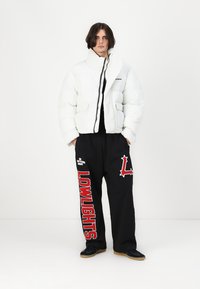Hvid pufferjakke med lynlås foran og høj krave, parret med sorte sweatpants med røde og hvide tekstgrafikker. Sorte sko fuldender looket.