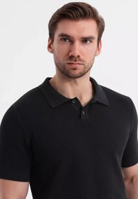 Musta lyhythihainen polo-paita, jossa on teksturoitu kuvio, kaulus ja kahden napin lista, joka tarjoaa vartalonmyötäisen muotoilun.