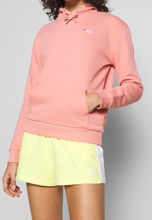 Person trägt einen pinken FILA-Hoodie mit Fronttasche und hellgelbe FILA-Shorts mit weißem Seitenstreifen vor grauem Hintergrund.