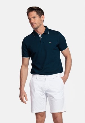 Marineblauw poloshirt met witte kraag en mouwaccenten, gedragen met witte shorts. Bevat een klein logo op de borst. Gladde katoenen stof.