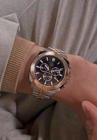 Sudraba un rozā zelta pulksteņi ar apaļu seju, melnu ciparnīcu, hronogrāfa funkcijām, teksturētu rāmi un saistītu metāla aproci.