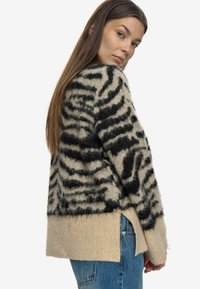 Uldblandet sweater med et sort og beige tigerstribemønster, ribbet kant og sideslidser for ekstra komfort. Model vist i profil.