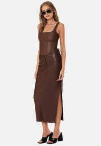 Abito in pelle marrone con corpetto strutturato, scollatura quadrata, spacchi laterali e silhouette aderente. La modella indossa sandali neri con tacco quadrato.