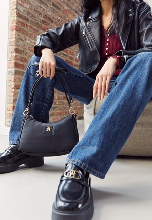 Osoba ubrana w czarną skórzaną kurtkę, czerwoną bluzkę, niebieskie jeansy, czarne mokasyny ze złotym detalem, trzymająca czarną torebkę z złotym logo, siedząca wewnątrz przy ceglanej ścianie.