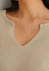 Gros plan d’une personne portant un haut en maille côtelée beige avec un large col en V et un détail en encoches près de la clavicule.