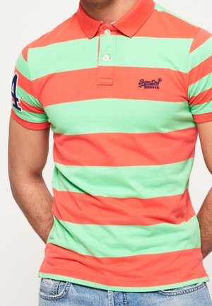 Mand iført kortærmet polo med brede vandrette striber i koral og mintgrøn, broderet logo på brystet, nummerlap på ærmet.