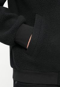 Il felpa nera in pile presenta una superficie testurizzata con polsini e orlo a coste. Tasca integrata nel design per funzionalità.