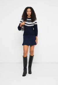 Dunkelblauer Pullover mit weißen geometrischen Mustern, kombiniert mit einem ausgestellt geschnittenen dunkelblauen Rock und kniehohen schwarzen Stiefeln. Hält eine kleine dunkle Handtasche.