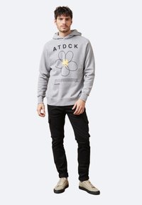 Sudadera gris con gráfico de flor negra y texto, combinada con pantalones cargo negros y zapatillas de color claro. El modelo está en una pose neutral.