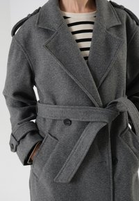 Cappotto grigio in misto lana con vita cinturata, ampi rever, epaulettes sulle spalle e tasche laterali. Presenta un tessuto texturizzato e chiusura con bottoni.