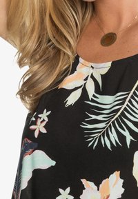 Femme blonde portant un haut noir à imprimé floral tropical pastel et un pendentif rond en bronze sur une chaîne en or.