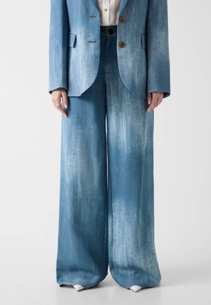 Frau trägt hellblaue, weit geschnittene Jeanshosen und einen passenden Blazer über einem weißen Hemd, steht vor einem einfarbigen Hintergrund.