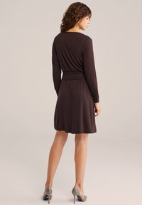 Vrouw met krullend haar, gekleed in een donkerbruine jurk met lange mouwen tot op de knieën en grijze hoge hakken, staand met haar rug naar de kijker en het hoofd iets zijwaarts gedraaid.