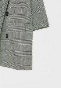 Manche et devant d'une veste à carreaux gris et noirs avec deux boutons noirs et une poche fendue horizontale sur fond blanc.