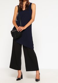 Navy sleeveless top with asymmetric hem, paired with black wide-leg trousers and black patent heels, holding a circular black handbag.