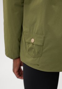 Gros plan d'une poche de veste verte avec rabat et bouton beige sur le côté gauche, portée sur un pantalon noir et un bras partiellement visible.