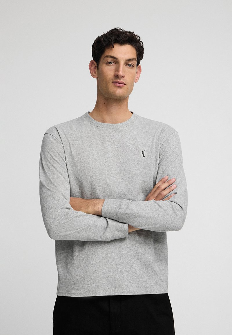 Szara koszulka z długim rękawem wykonana z miękkiego materiału, z okrągłym dekoltem i małym czarnym logo na lewej piersi. Prosty krój u dołu.