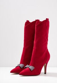 Bottes de cheville en suede rouge avec un bout pointu, un talon haut et un accent décoratif en nœud à carreaux à l'avant. Design élégant et ajusté.