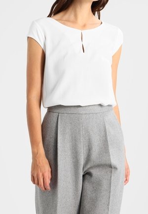 Frau trägt eine weiße Kurzarmbluse mit kleinem Vorderschlitz, in eine hoch taillierte graue plissierte Hose gesteckt.