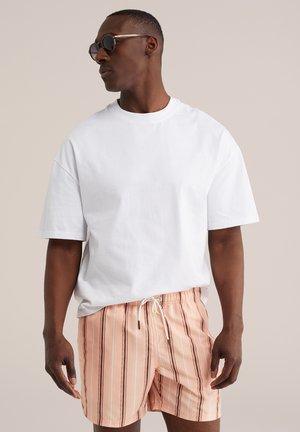 Férfi kerek napszemüveget visel, fehér túlméretezett pólót és barackszínű csíkos, megkötős rövidnadrágot, bézs háttér előtt áll.