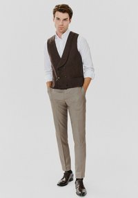 Brązowa kamizelka dwurzędowa na białej koszuli, w połączeniu z jasnobeżowymi spodniami w paski i brązowymi eleganckimi butami.