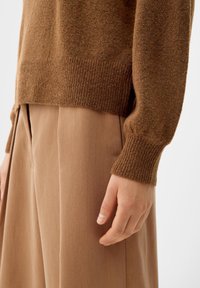 Personne portant un pull en tricot marron avec des poignets côtelés et un pantalon large plissé beige, la main détendue à leurs côtés.