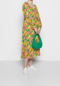 Blommig midiklänning i livfulla nyanser av gult, rosa och grönt; rund halsringning; långa ärmar. Håller en grön, böjd handväska. Vita sneakers synliga.