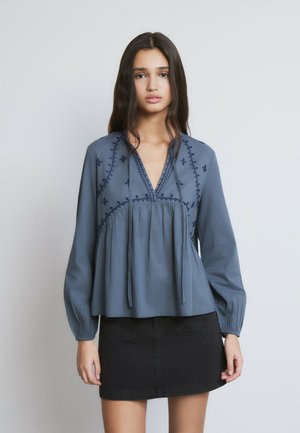 Joven con cabello largo y oscuro, vestida con una blusa azul bordada de manga larga y una minifalda negra de mezclilla, posando frente a un fondo liso.