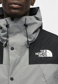 Γκρι και μαύρο σακάκι με ψηλό γιακά, ρυθμιζόμενα κορδόνια, κουμπιά με κλικ και κεντημένο λογότυπο στο στήθος.