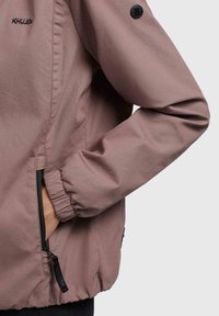 Helle rosa Jacke aus glattem Stoff. Verfügt über elastische Bündchen, Seitentaschen mit schwarzen Reißverschlüssen und einem Logopatch am Ärmel.