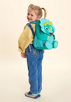 Smiling young girl with pigtails wearing yellow sweatshirt, blue overalls, and a teal dinosaur-themed backpack facing sideways.