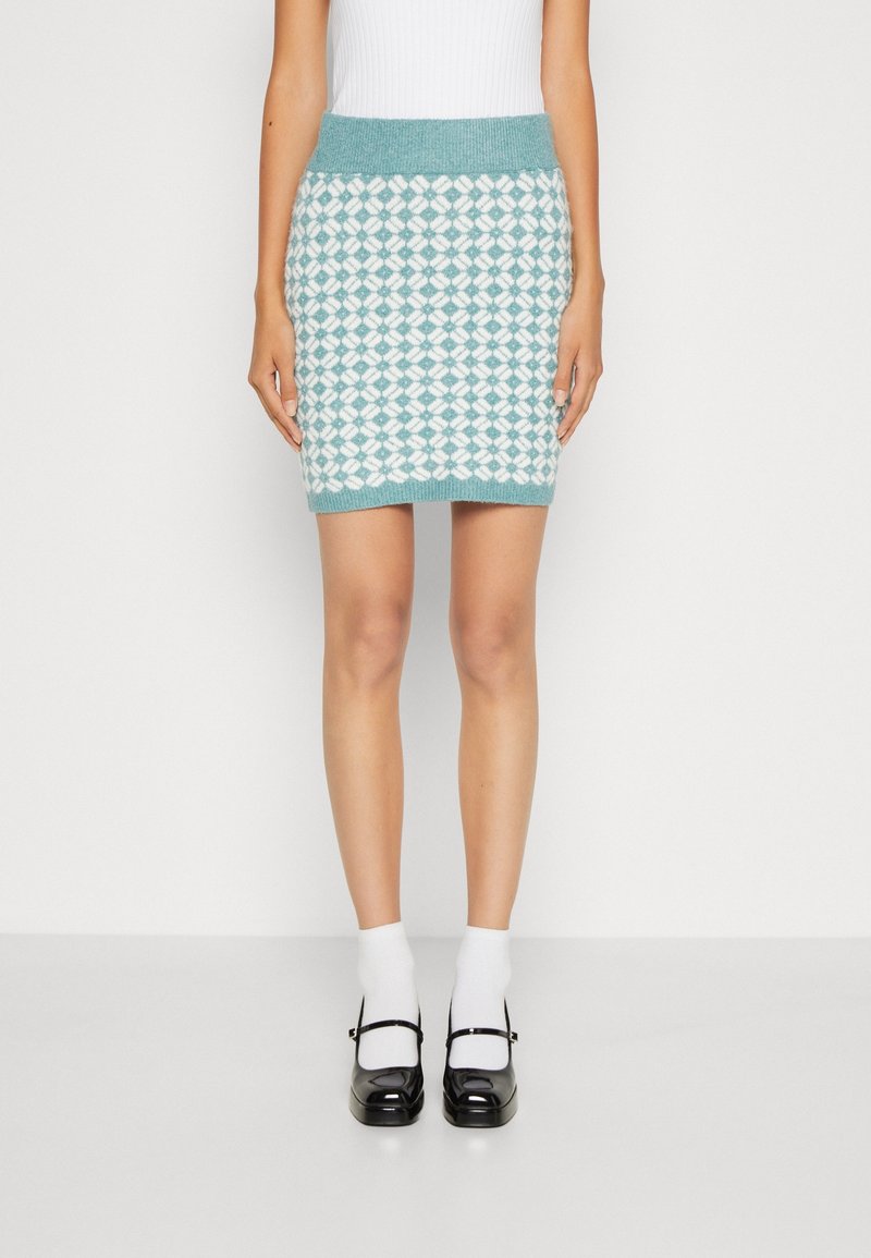Femme portant une mini-jupe tricotée à motif vert sarcelle et blanc, des chaussettes blanches et des chaussures Mary Jane noires, debout devant un fond uni.