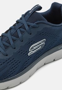 Navy blue athletic shoe with mesh upper, textured detailing, and grey sole. Features logo on the side and flat laces.