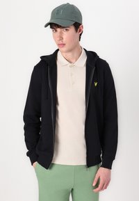Hanorac negru cu fermoar, cu buzunare laterale, purtat peste un tricou polo bej, asociat cu pantaloni de trening verzi și o șapcă gri.
