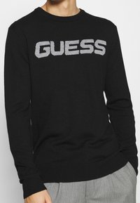 Schwarzer Strickpullover mit Rundhalsausschnitt, der ein auffälliges graues "GUESS"-Logo auf der Vorderseite trägt. Gerippte Bündchen und Saum. Strukturiertem Stoff.