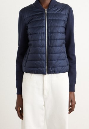 Personne portant une veste matelassée bleue marine zippée avec des manches tricotées et un pantalon blanc taille haute, debout devant un fond uni.