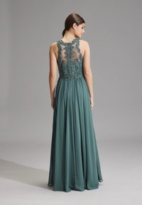 Lungo vestito teal con un corpetto in pizzo trasparente decorato con ricami floreali. La gonna fluente cade da una vita aderente, creando un movimento elegante.