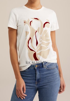 Persoon draagt een wit T-shirt met korte mouwen met beige en donkerrood abstract bladprint en een blauwe spijkerbroek tegen een neutrale achtergrond.