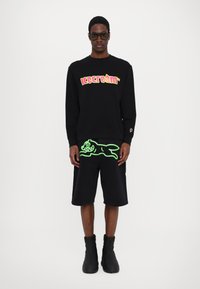 Svart hettegenser med flerfarget "is-krem" grafikk. Svart shorts med neongrønn kattemotiv. Svart ankelstøvler.